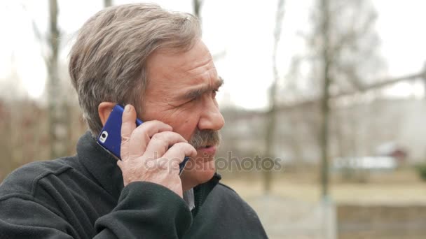 Een oudere man met een snor rookt en gesprekken over de telefoon in de straat. De oude huizen liggen achter ons. De zwarte jas. — Stockvideo