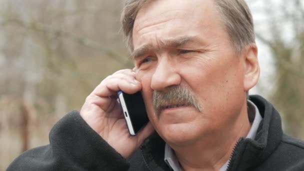 Un anciano con bigote fuma y habla por teléfono en la calle. Las viejas casas están detrás de nosotros. La chaqueta negra . — Vídeo de stock
