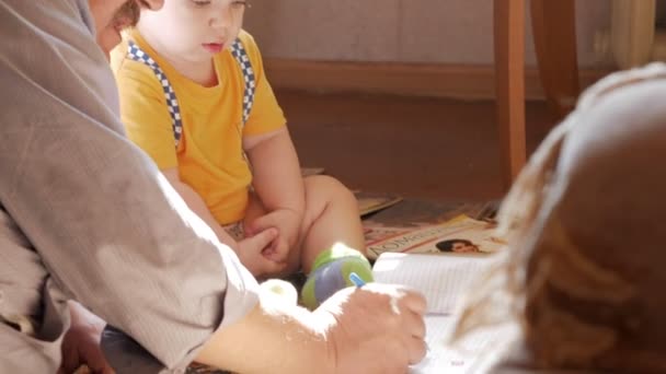 Grand-père et petit-fils peignent une maison par terre sur une feuille de papier. La lumière du soleil illumine l'image . — Video