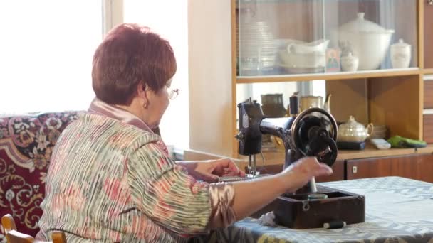 Mujer adulta cose en la vieja máquina de coser manual. Las gafas están vestidas y la tela gris está cosida . — Vídeos de Stock