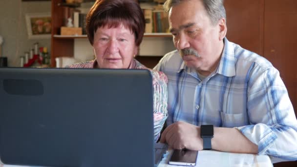 Ένα ηλικιωμένο ζευγάρι κάθεται στο σπίτι με το laptop. Μια γυναίκα διαβάζει ειδήσεις, ένας άνθρωπος με μουστάκι που κάθεται δίπλα του και συνομιλίες — Αρχείο Βίντεο