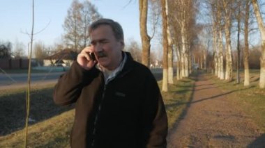 Yaşlı bir adam parkta elini kolunu sallaya dönüyor, dumanlar ve cep telefonundan konuşuyor. Yakınlarda otomobil bir yoldur