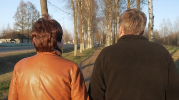Loving elderly couple walking in the park at sunset. Nearby there is a road. A woman in a brown leather jacket. The man is dressed in a black sweater. — Stock Video
