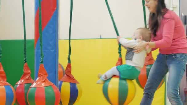 Mom rolls a pretty kid on an attraction with a hanging swing. The boy holds on to the ropes. — Stock Video