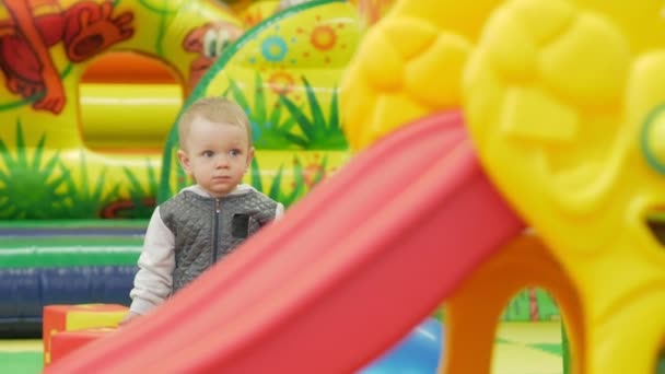 Um garoto bonito está parado perto de um slide vermelho em um parque de diversões — Vídeo de Stock
