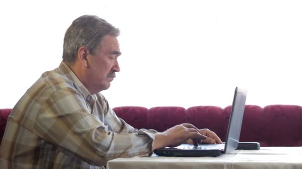 Un anciano con bigote se sienta detrás de un portátil y resuelve problemas. Mira seriamente al monitor. — Vídeo de stock