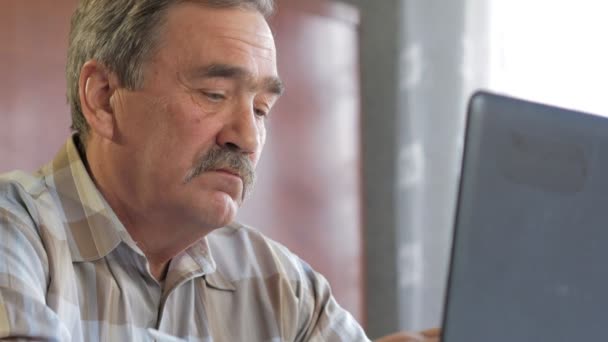 Un anciano con bigote se sienta detrás de un portátil y resuelve problemas. Mira seriamente al monitor. — Vídeos de Stock