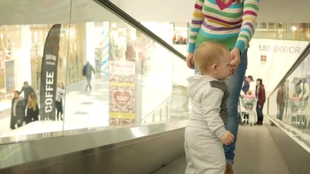 A cute blond boy of two years with his mother climbs the escalator in a large shopping center. The kid looks all around — Stock Video