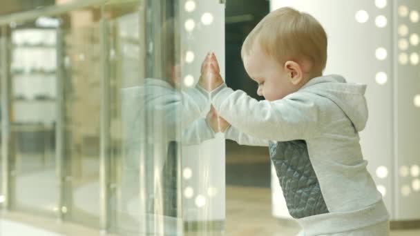 Egy aranyos szőke fiú kétéves fontolgatja egy boltban, az üvegen keresztül. A gyerek gondosan megvizsgálja a belsejében a boltban — Stock videók
