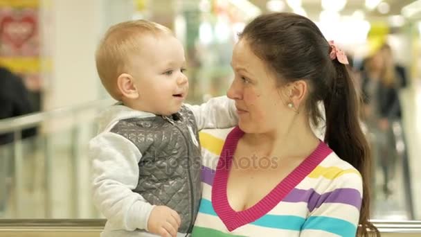 A mãe tem um lindo filho loiro nos braços no centro comercial. Contra o pano de fundo as pessoas vão às compras — Vídeo de Stock