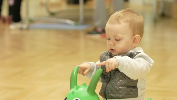 Aranyos szőke fiú két éves ugrás egy felfújható ló a plázában. Gyerek rohangál, a háttérben. — Stock videók