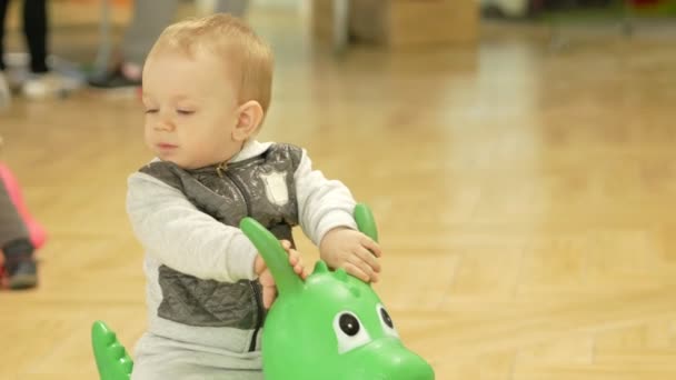 Lindo chico rubio de dos años saltando sobre un caballo inflable en el centro comercial. Los niños están corriendo en el fondo . — Vídeo de stock