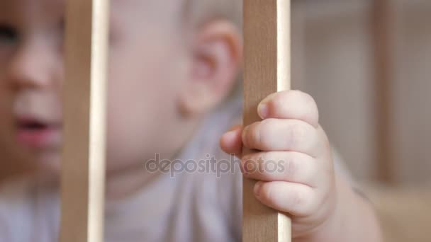 Bonito bebê loiro mostra jogando em um berço de bebê. Em foco, a mão segura cercas — Vídeo de Stock
