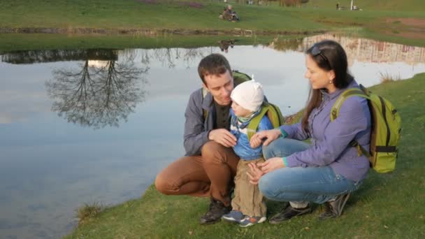 Uma jovem família ativa com um bebê senta-se junto ao lago no parque e fala uns com os outros — Vídeo de Stock