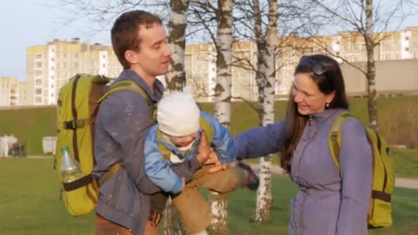 Familia activa con el bebé camina en el parque en la primavera. Hierba verde y lago cerca. Mochilas verdes de diferentes tamaños — Vídeos de Stock