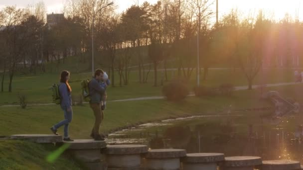 Actieve familie met de baby gaat een mooie brug over de rivier bij zonsondergang. De zon schijnt in het frame — Stockvideo