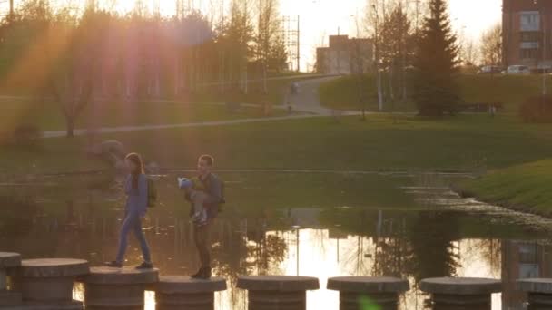 Aktivní rodina s dítětem předává krásný most přes řeku při západu slunce. Slunce svítí v rámu — Stock video