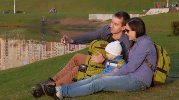 Una giovane famiglia attiva con un bambino si siede vicino al lago nel parco e parla tra di loro. — Video Stock