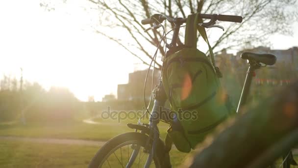 夕暮れ時に公園で折りたたみ自転車は立っています。それに掛けるグリーン バックパック — ストック動画