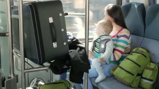 Мама и ребенок сидят в движущемся поезде возле окна. Чемодан и рюкзаки расставлены рядом с молодой семьей. Сын на ногах у матери — стоковое видео