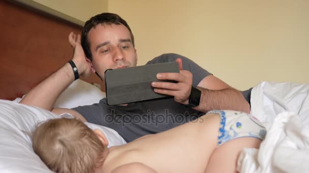 Far läser nyheterna på tabletten nära det sovande barnet. En pojke som vilar i förgrunden av ett hus på en säng — Stockvideo