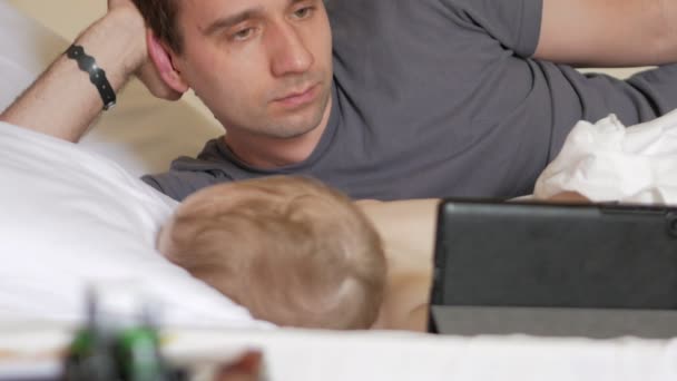 Батько читає новини на планшеті біля сплячого малюка. Хлопчик відпочиває на передньому плані будинку на ліжку — стокове відео