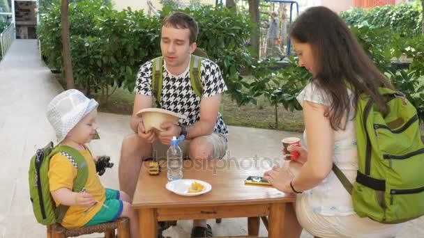 Giovane famiglia felice con un bambino che mangia all'aria aperta in un caffè. Tutti sono seduti su piccole sedie di legno — Video Stock