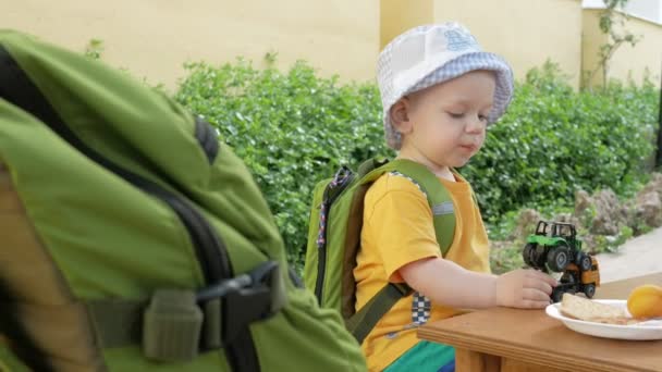 Söt liten äter utomhus på ett café. Han leker med bilar och äter kakor med vatten — Stockvideo