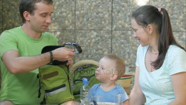 Una familia joven con un niño está midiendo las gafas de sol en un bebé. Mamá papá y bebé están sentados en la cafetería del parque — Vídeos de Stock