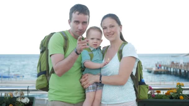 Молодая семья с малышом, смотрящим в камеру и улыбающимся. У всех рюкзаки на спине. Море и пляж позади . — стоковое видео