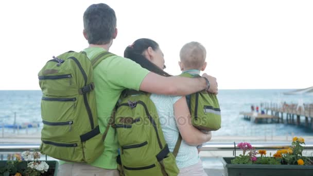 Familia joven con un bebé mirando al mar y abrazándose. Todo el mundo tiene mochilas en la espalda. Playa y gente — Vídeos de Stock