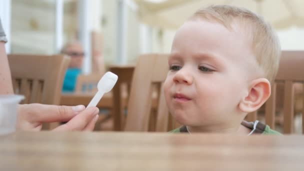 Mère nourrit son jeune fils de crème glacée à partir d'une cuillère. Ils sont assis dans un café de rue. Le garçon aime vraiment la délicatesse . — Video