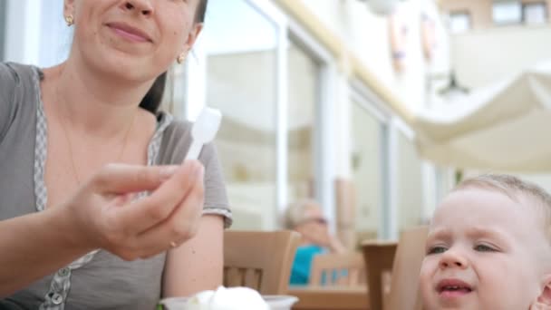 Mère nourrit son jeune fils de crème glacée à partir d'une cuillère. Ils sont assis dans un café de rue. Le garçon aime vraiment la délicatesse . — Video