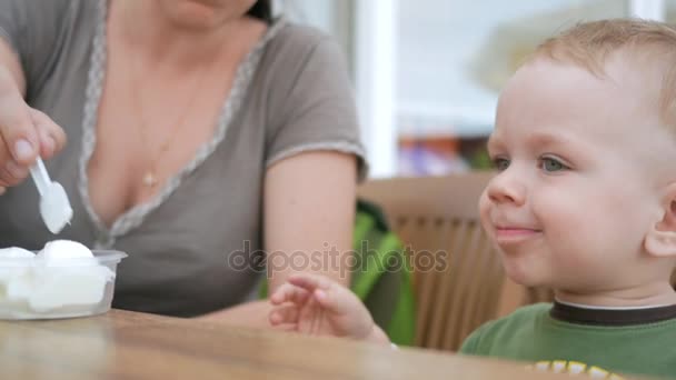 A mãe alimenta seu filho com sorvete de uma colher. Eles estão sentados em um café de rua. O rapaz gosta mesmo da iguaria. . — Vídeo de Stock