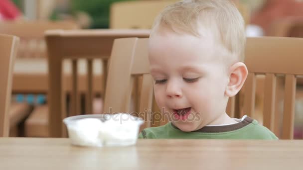 Mother feeds her young son with ice cream from a spoon. They are sitting in a street cafe. The boy really likes the delicacy. — Stock Video