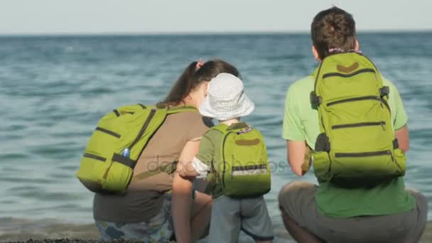 Молодая семья с ребенком, смотрящим на море и обнимающимся. У всех рюкзаки на спине. Пляж и люди — стоковое видео