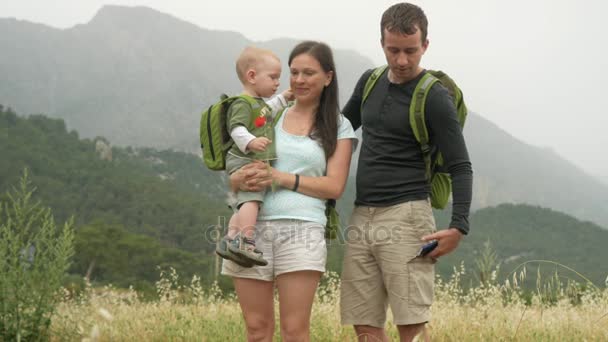 Una joven familia de viajeros con un niño pequeño está mirando la cámara cerca de las hermosas montañas. Todo el mundo sonríe y juega con su hijo . — Vídeo de stock