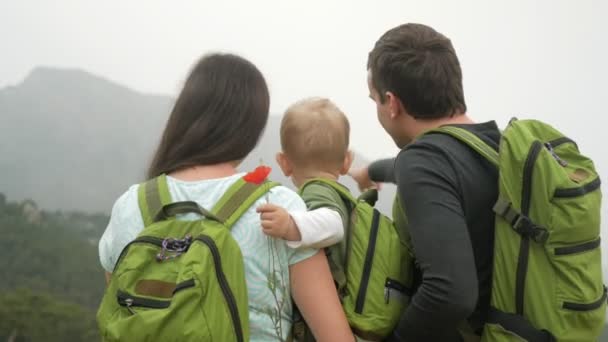 Mladá rodina cestující s dítětem při pohledu zpět na horách. Každý má stejné zelené batohy. — Stock video