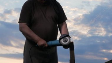 Bir erkekte yaş bir metal açılı taşlama günbatımında keser. İşçi arkasında güzel gökyüzü