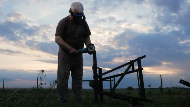 Мужчина в возрасте отрезает металлическую угловую мельницу на закате. Красивое небо за рабочим — стоковое видео