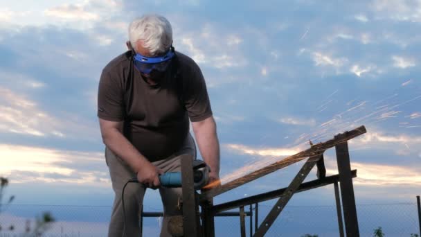 Een man in de leeftijd afsnijdt een metalen Haakse slijper bij zonsondergang. Mooie hemel achter de werknemer — Stockvideo