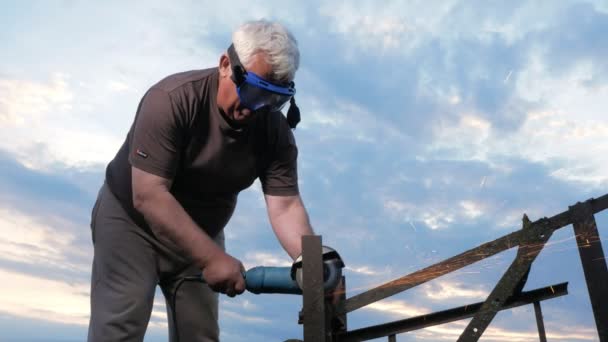 Een man in de leeftijd afsnijdt een metalen Haakse slijper bij zonsondergang. Mooie hemel achter de werknemer — Stockvideo