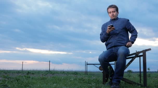Ein junger Mann checkt bei Sonnenuntergang die Nachrichten am Telefon. dahinter ein Feld mit grünem Gras. schöner Himmel. — Stockvideo