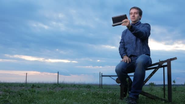 Un joven está sentado y hablando en la tableta al atardecer. Un campo de hierba verde detrás. Hermoso cielo . — Vídeos de Stock