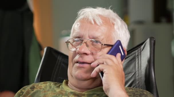 En man vid en ålder av sitter i en skinnfåtölj och pratar på sin mobiltelefon. Klädd Glasögon, grå hår — Stockvideo