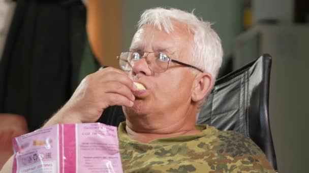 Hombre mayor comiendo palitos de maíz y viendo televisión. Se sienta en un sillón de cuero con gafas — Vídeo de stock