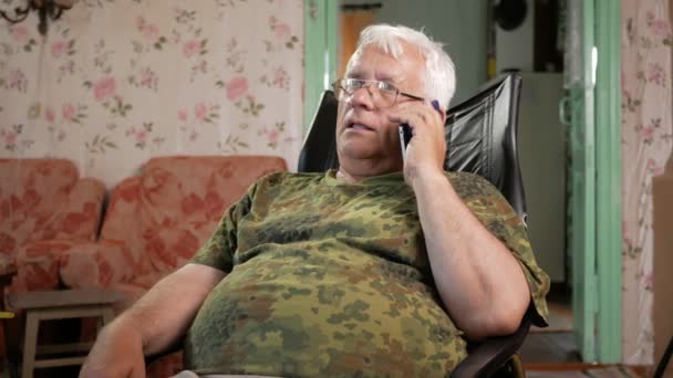 Un homme à l'âge de s'asseoir sur une chaise en cuir et de parler sur son téléphone portable. Lunettes habillées, cheveux gris — Video