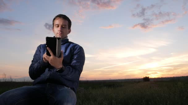 Молодий чоловік сидить на заході сонця і перевіряє повідомлення, щоб перевірити повідомлення на планшеті. Прекрасне небо позаду . — стокове відео