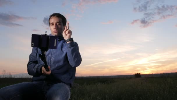 Ein junger Mann sitzt bei Sonnenuntergang und macht ein Selfie mit seinem Handy. schöner Himmel dahinter. — Stockvideo