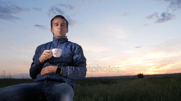 Un jeune homme boit une boisson chaude dans une tasse au coucher du soleil. Beau ciel derrière . — Video